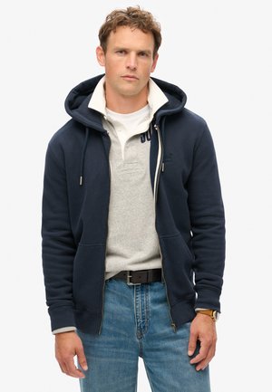 Marineblå zip-up hoodie med snoretræk, lagdelt over en grå polo. Båret med lyseblå jeans og et sort bælte, har en struktureret stof.