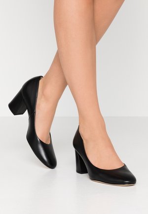 Kvinde iført sorte lukkede pumps med blokhæl i glat læderfinish, stående på en ensfarvet hvid overflade.