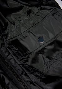 Close-up of black shiny fabric with a zippered seam and a small pocket secured by a metallic snap button.