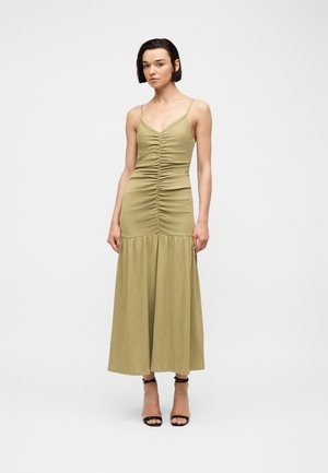 Femme portant une robe midi sans manches froncée vert olive avec fines bretelles et sandales noires à talons hauts, debout sur un fond uni.