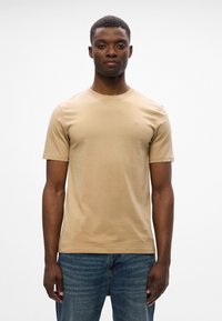 Camiseta beige de manga corta con cuello redondo y un sutil detalle de logo, combinada con jeans de denim azul. Textura suave y diseño ajustado.