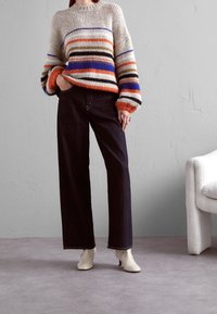 Pull en maille à rayures multicolores orange, bleu, vert et crème. Associé à un jean large foncé et des bottines à talons beiges.