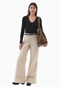 Donna che indossa un top a maniche lunghe nero con scollo a V, pantaloni beige a gamba larga con risvolto, cintura nera, scarpe nere e che porta una borsa a tracolla con stampa leopardata.