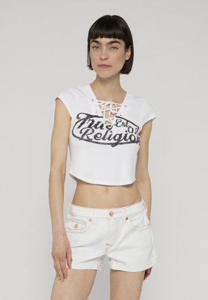 Žena s krátkými tmavými vlasy, která má na sobě bílý krátký top s šněrováním a nápisem a bílé džínové kraťasy, stojící před jednoduchým pozadím.