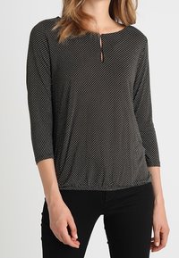 Schwarzes Oberteil mit weißen Punkten, runder Ausschnitt, Dreiviertelärmel und einem kleinen Schlüsselriemendetail am Hals. Weicher, dehnbarer Stoff.