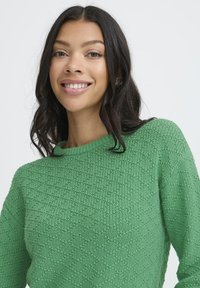 Mujer joven con cabello largo y oscuro sonriendo, vistiendo un suéter de punto verde texturizado, contra un fondo claro y liso.