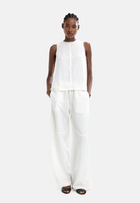 Biała bluzka bez rękawów z surowymi krawędziami, w połączeniu z luźnymi, przezroczystymi białymi spodniami. Modelka nosi czarne sandały na odkrytych palcach.