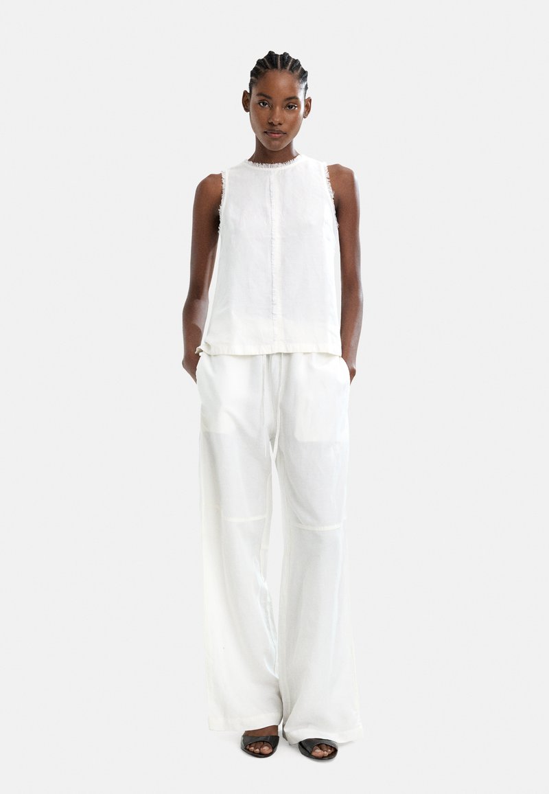 Biała bluzka bez rękawów z surowymi krawędziami, w połączeniu z luźnymi, przezroczystymi białymi spodniami. Modelka nosi czarne sandały na odkrytych palcach.