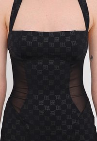 Rochie neagră, ajustată, cu un model ce prezintă un motiv de logo gri. Panouri laterale din plasă transparentă, decolteu pătrat și bretele late.