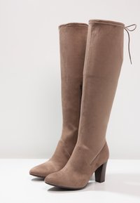 Botas de ante marrón hasta la rodilla con punta afilada, tacón alto y cremallera lateral; cuentan con un detalle de encaje en la parte superior.