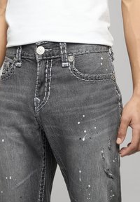 Grijze distressed jeans met spetterpatronen en lichte stiksels. Voorzien van een metalen knoop en klinknagels, benadrukt door een gestructureerde denimafwerking.
