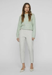 Un pull en maille vert clair avec des motifs perforés, associé à un pantalon sur mesure gris clair et des escarpins beiges à bout pointu.