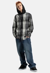Camisa de manga larga a cuadros en negro y gris, con capucha negra, combinada con jeans azules holgados y zapatillas negras sobre un fondo neutro.