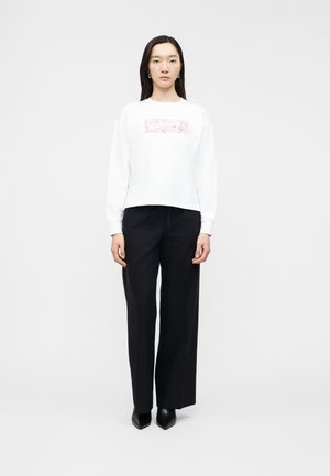 Γυναίκα όρθια, φοράει λευκό τοπ με μακριά μανίκια και ρόζ γραφικό, μαύρο παντελόνι φαρδύ και μαύρα παπούτσια με τακούνι, μπροστά σε λευκό φόντο.