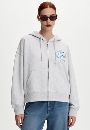 Femme aux cheveux roux et lunettes de soleil foncées portant un sweat à capuche gris clair zippé avec un motif requin coloré et un jean bleu.