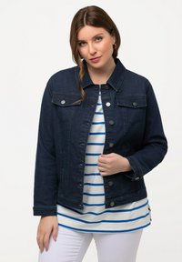 Giacca in denim scuro con design a bottoni, due tasche frontali e colletto. Indossata sopra una maglietta a righe bianca e blu.