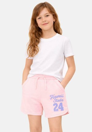 Jong meisje met lang rood haar, gekleed in een wit T-shirt en roze shorts met 'Hawaii State 24' in het blauw erop gedrukt, handen in de zakken.