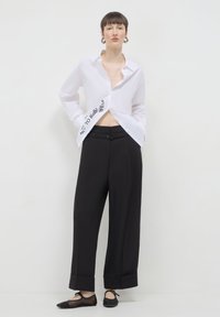 Camicia bianca cropped con chiusura a bottoni e testo stampato sul lato, abbinata a pantaloni neri a vita alta e gamba larga e ballerine nere.