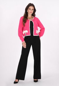 Cárdigan de punto rosa con ribetes blancos y detalles de lazos, combinado con pantalones anchos negros. Textura suave, diseño casual.