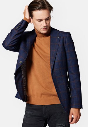 Granatowy blazer w brązową kratę, jednozapięciowy fason z wzorzystym wnętrzem, połączony z rdzawej barwy swetrem z golfem i przetartymi dżinsami.