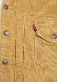 Tissu en velours côtelé jaune moutarde avec des côtes verticales. Comprend des boutons en métal et une poche avec une étiquette rouge sur le côté.