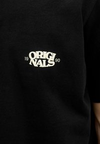 Černé tričko s krátkým rukávem se bílým potiskem „ORIGINALS 1990“ na levé části hrudi, vyrobené z hladkého bavlněného materiálu.