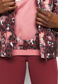Jaguarprint jakke med lyserøde detaljer, med lynlås og trykknapper. Underneath en lys rosa skjorte og mørk rosa tætsiddende bukser.