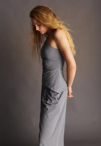 Femme aux longs cheveux blonds regardant vers le bas, portant une robe grise sans manches ajustée, debout devant un fond neutre uni.