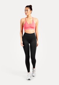 Kvinna som bär en rosa sportbehå, svarta högmidjade leggings och vita sneakers, stående och tittar åt vänster mot en vit bakgrund.