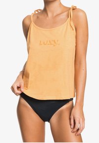 Orange ärmlös topp med knytband, med en urblekt logga. Kombinerad med svarta badbyxor som visar en avslappnad passform.