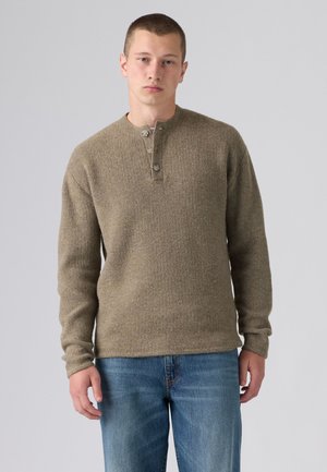 Jeune homme portant un pull en maille marron à manches longues avec des boutons et un jean bleu, debout devant un fond clair uni.