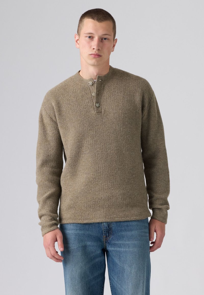 Jeune homme portant un pull en maille marron à manches longues avec des boutons et un jean bleu, debout devant un fond clair uni.