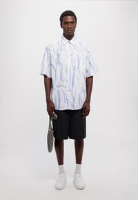 Cămașă cu mânecă scurtă, albă, cu un model abstract albastru, purtată cu pantaloni scurți negri și teniși albi, ținând o geantă gri.