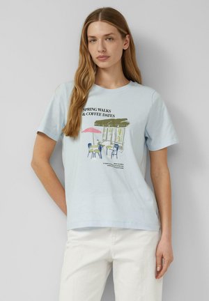 Kobieta z długimi włosami nosi jasnoniebieską koszulkę z nadrukiem kawiarni na świeżym powietrzu i napisem „Spring Walks & Coffee Dates”, w połączeniu z białymi spodniami.