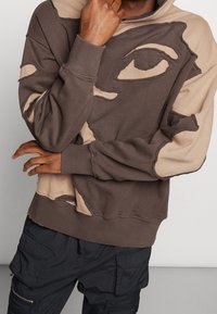 Felpa marrone con accenti beige, caratterizzata da un design di volto astratto, dettagli usurati e una vestibilità rilassata. Abbinata a pantaloni cargo neri.