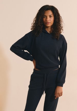 Vrouw met krullend haar die een donker geribde lange mouwen hoodie en bijpassende broek draagt, staande met één hand op de heup tegen een beige achtergrond.