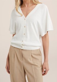 Cardigan bianco con maniche corte e scollo a V, dotato di chiusura con bottoni. Realizzato in tessuto morbido, presenta una vita fitted e una trama delicata.