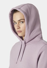 Světle fialová mikina s kapucí na šňůrku, jemnou texturou a bočními švy. Disponuje volným střihem a minimalistickým designem.