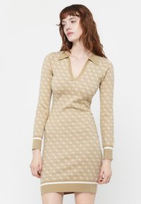 Rochie tricotată bej cu decolteu în V, maneci lungi și guler. Prezintă un model geometric alb și benzi contrastante la manșete și la tiv.