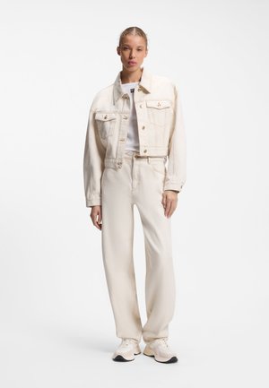 Mujer con chaqueta vaquera color crema y vaqueros de talle alto y pierna ancha a juego con zapatillas blancas sobre fondo liso.