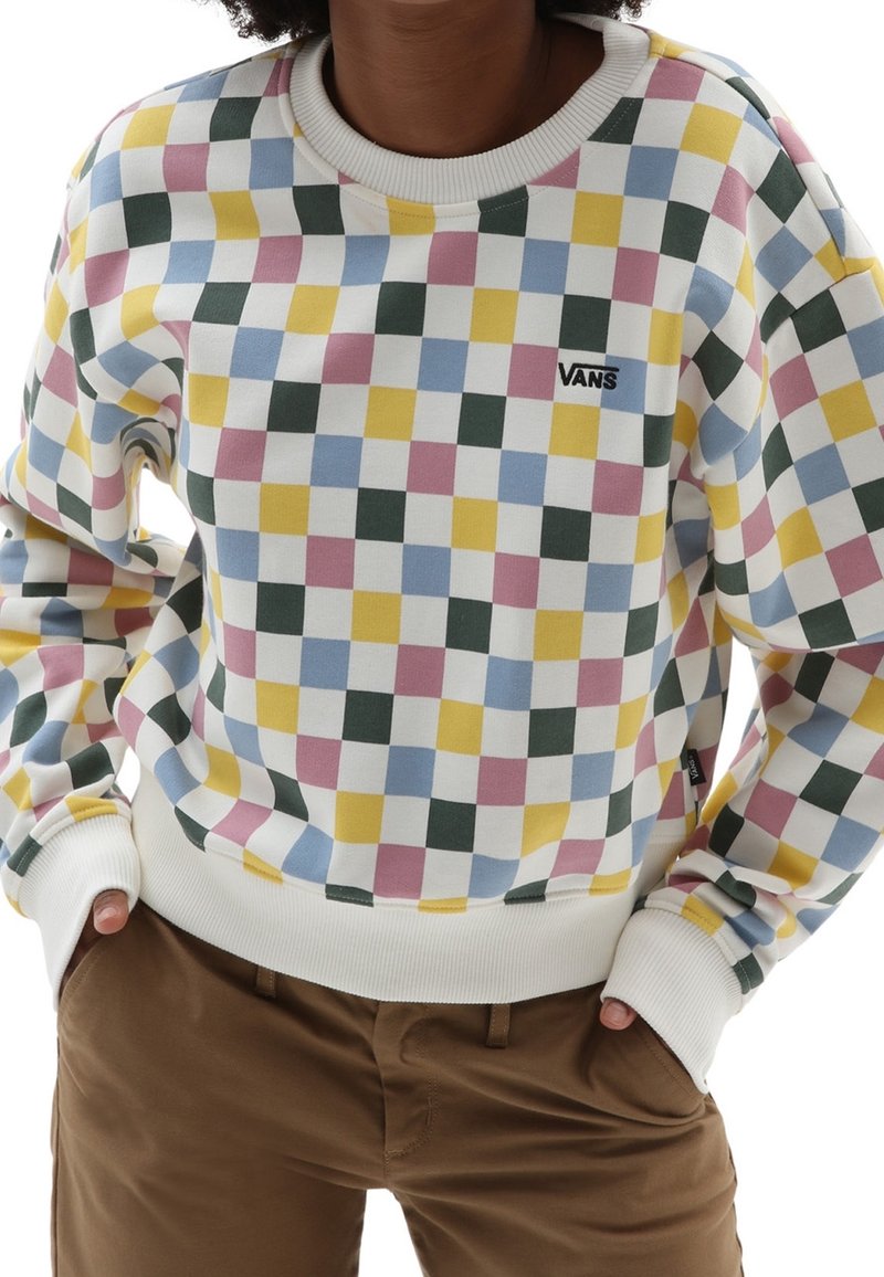 Personne portant un sweat Vans à carreaux multicolores avec des poignets blancs et un pantalon marron, les mains dans les poches.