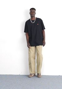 Schwarzes T-Shirt mit kurzen Ärmeln und kleinem orangefarbenem Logo, beiges, gerades Hosenmodell und braune Sneaker mit weißen Streifen. Einfaches Design.