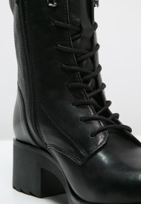 Bottine en cuir noir avec talon épais, lacets sur l'avant, fermeture éclair latérale et coutures détaillées sur la surface supérieure lisse.