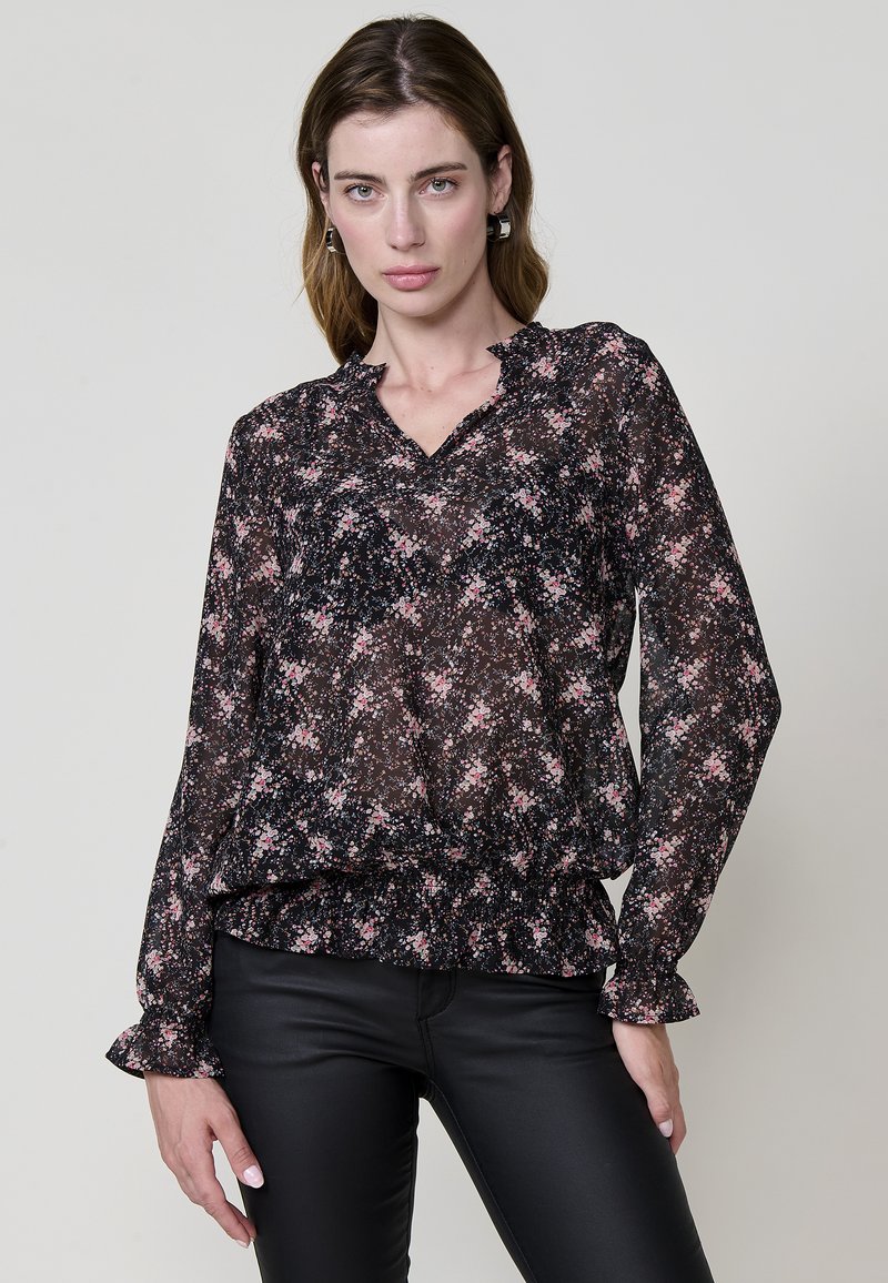 Sort blomstret bluse med lange ærmer, flæser på manchetterne, v-udskæring og samlet kant; har pink, hvide og grønne blomsterprint på gennemsigtigt stof.