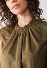 Olivgrön ärmlös topp med en samlad necklinje, tillverkad av mjukt tyg med en subtil drapering och slät yta.