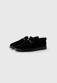 Svarte loafers med overdel i semsket skinn, myk fôr av pels, dekorative sømmer og metallakenter på fronten, og med flat gummisåle.