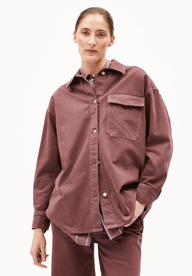 Frau mit nach hinten gegeltem Haar, die ein lockeres, mauvefarbenes Hemd mit Knöpfen und einer Brusttasche über einem gestreiften Oberteil und passenden Hosen trägt und mit einer Hand in der Tasche steht.