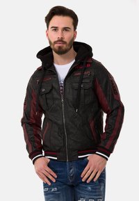 Schwarzlederjacke mit maroonfarbigen Akzenten, Reißverschlusstaschen, gestreiften Manschetten und einer abnehmbaren schwarzen Kapuze. Textdetails an den Ärmeln sind sichtbar.