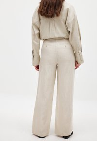 Beige Leinenoutfit mit einem lockeren Hemd und weit geschnittenen Hosen, versehen mit Knopfdetails an den Bündchen und Gesäßtaschen, welches eine lässige Silhouette erzeugt.