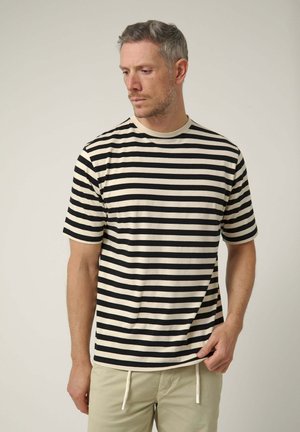 Hombre con el cabello corto y gris, vistiendo una camiseta de rayas horizontales en blanco y negro y pantalones beige, mirando hacia abajo a su izquierda.